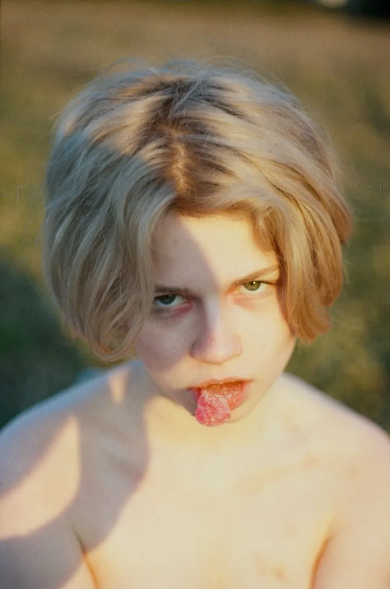 Film portrait in late sun: a blonde with a short bob glares up at the lens, a sugared red sweet on her tongue, bare shoulders