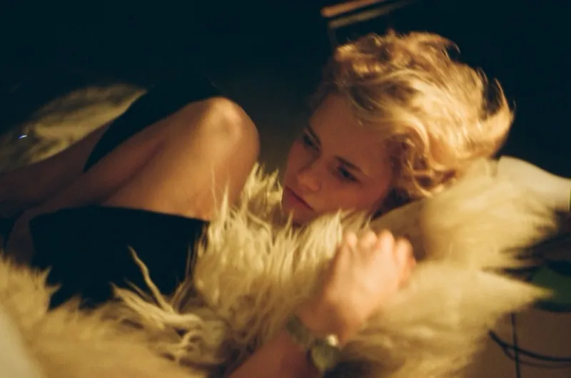 Low-light film frame: a blonde with tousled curls lies in a shaggy white fur, gazing aside, a hand resting on the fur