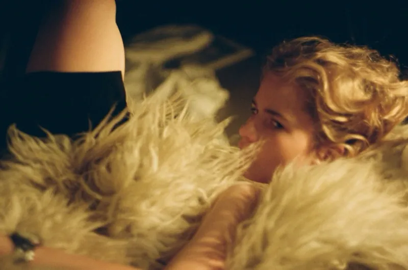 Warm close frame: the same blonde peers over a cloud of white fur, eyes turned to the side, a figure in black blurred beside her