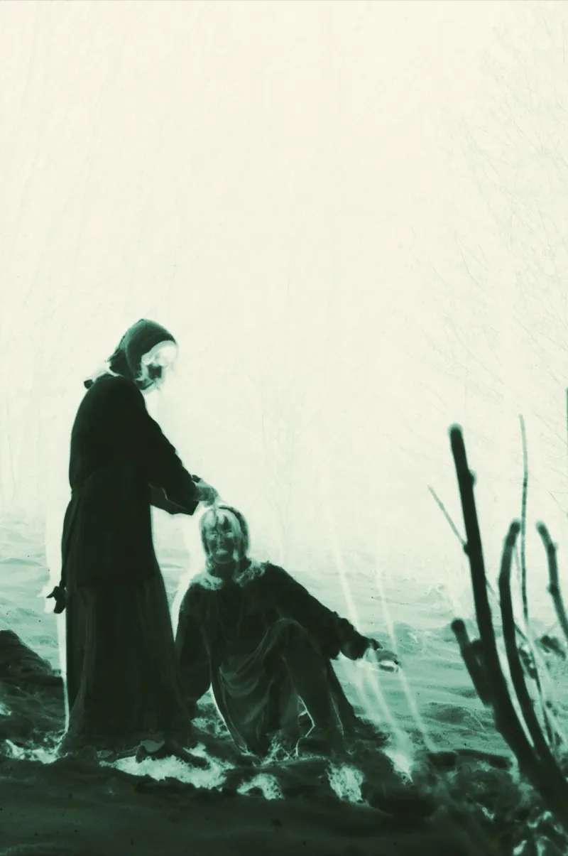 Green-toned negative: a figure in a headscarf holds her hands over the head of a woman sitting on the ground, mist and bare branches behind