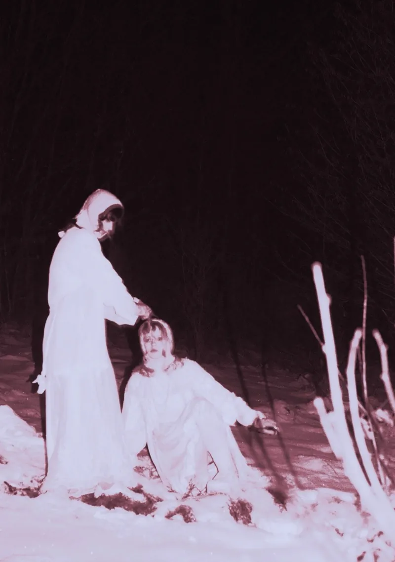 Pink-toned night scene: a figure in a white robe holds her hands over the head of a woman sitting in the snow, flash-lit branches beside them