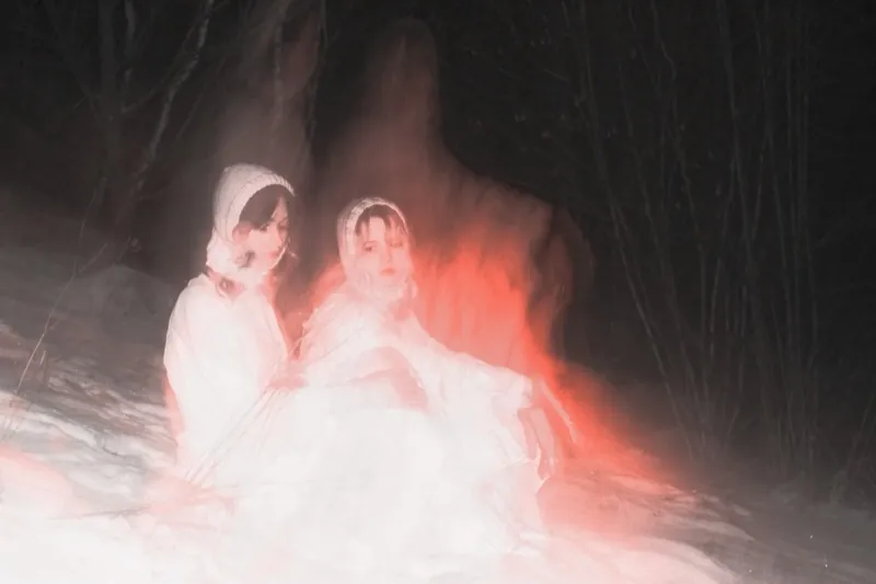 Two women in white bonnets sit in the snow, behind them a blurred red-orange streak of light shaped like a figure