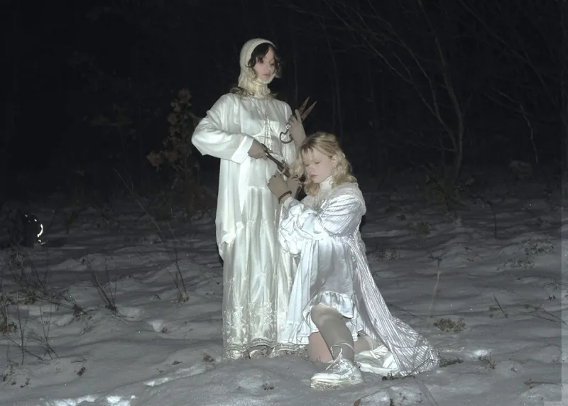 Snowy forest at night: a woman in a white dress and knitted bonnet holds old shears, a kneeling blonde in a satin dress rests her head against her side