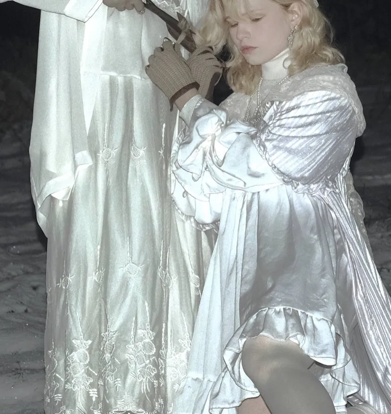 Close-up: a fair-haired woman in a puff-sleeved satin dress kneels by a figure in an embroidered dress, knit-gloved hands on the shears
