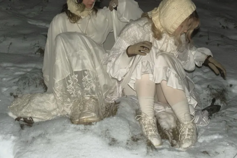 The same moment in a warmer tone: two women in white rest on the snowy ground, old shears lying beside them