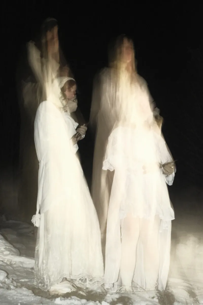 Long exposure: two figures in white dresses stand on the snow at night, blurred translucent silhouettes behind them