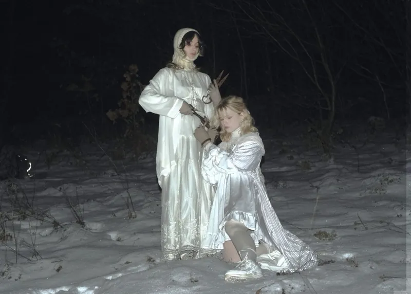 Snowy forest at night: a dark-haired woman in a white satin dress and knitted bonnet holds rusty shears over a kneeling blonde in a striped satin gown, who grips the blades in knit gloves