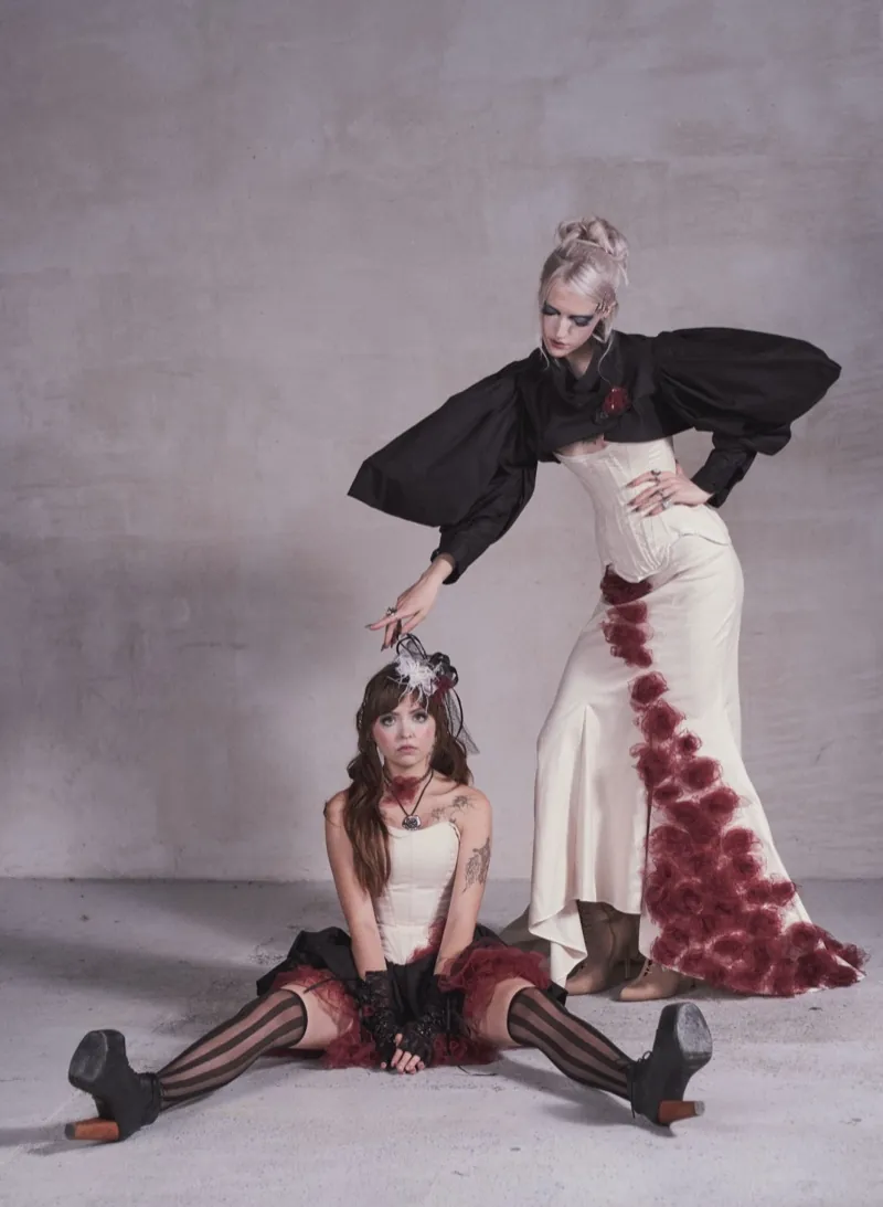Two models in the studio: a brunette in a cream corset and striped stockings sits on the concrete floor, a blonde in a puff-sleeved black bolero and a cream gown with burgundy roses leans over her