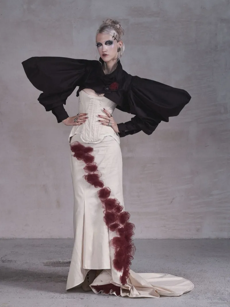 A blonde in heavy dark makeup stands with hands on hips: a black bolero with huge sleeves, a cream corset and a gown cut diagonally by a band of burgundy roses