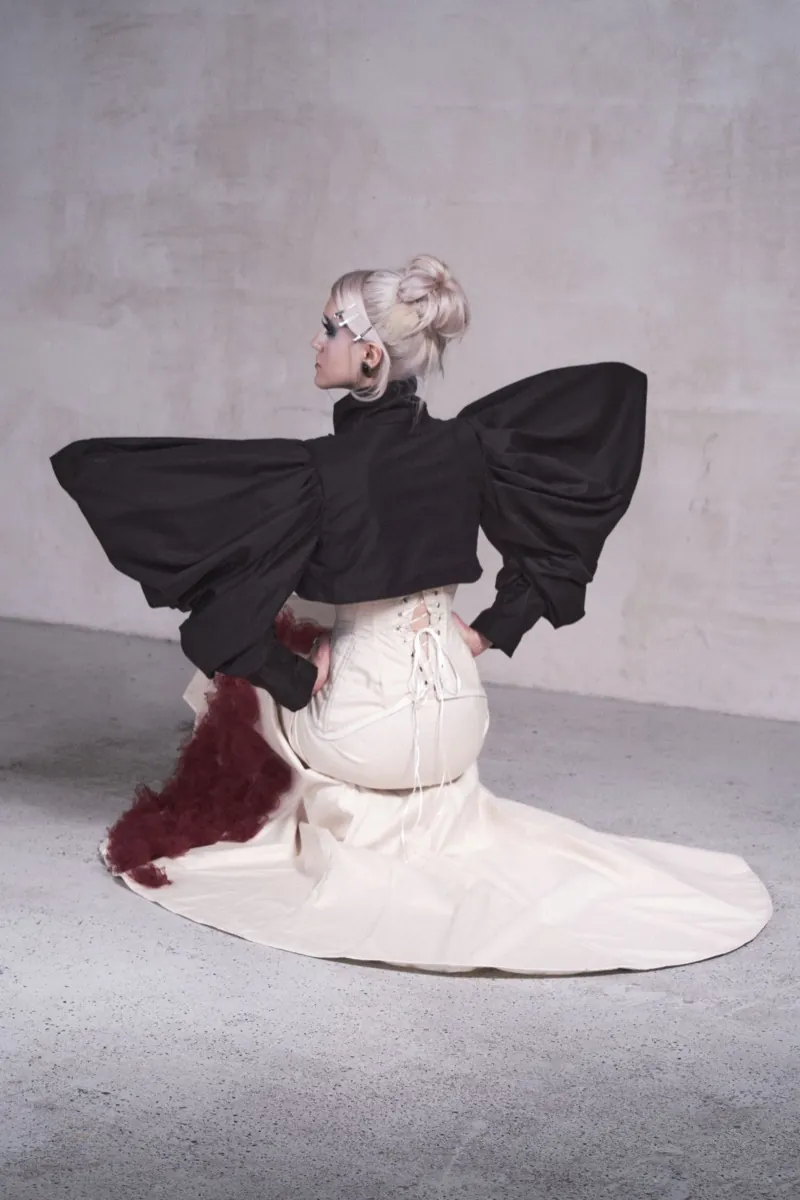 A model kneels with her back to the camera, black bolero sleeves spread like wings, a laced corset and a train of burgundy roses on the concrete