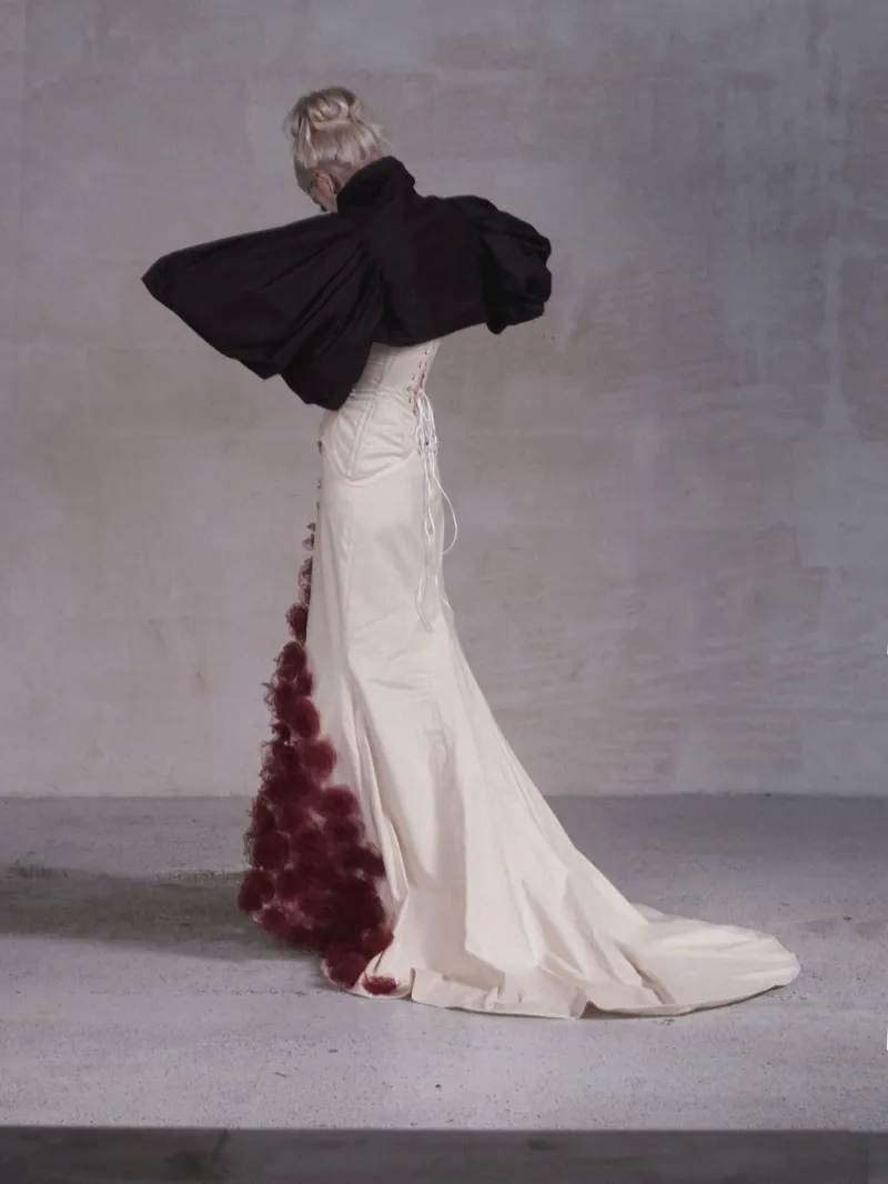 A model stands in profile with her head bowed, black puffed sleeves over a cream gown with a long train and a cascade of burgundy roses