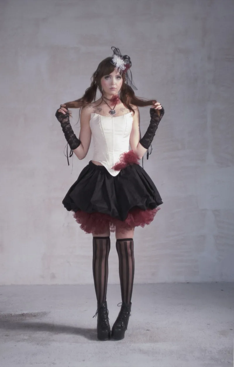 A brunette faces the camera in a cream corset, a black puffed skirt with burgundy tulle and striped stockings, holding strands of her hair