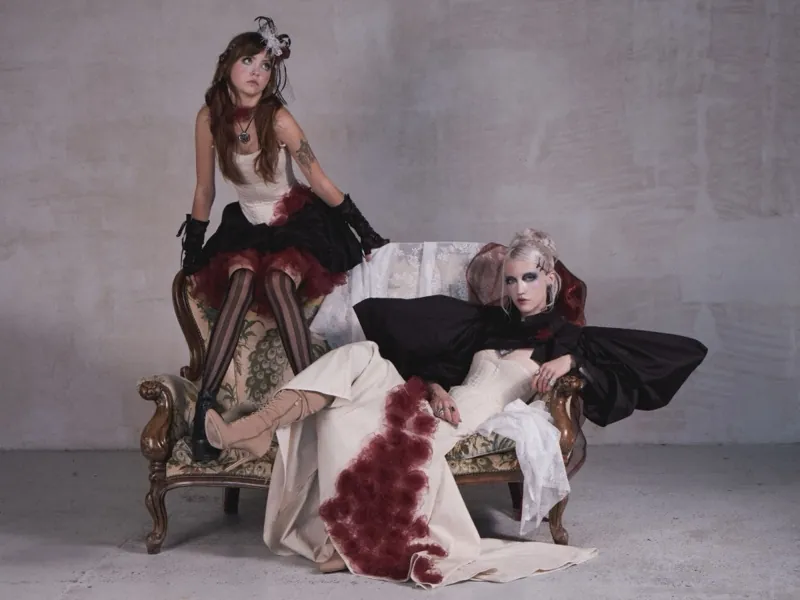 A brunette sits on the arm of an antique peacock-upholstered sofa, beside her a blonde reclines in a cream gown with burgundy roses and outspread black sleeves
