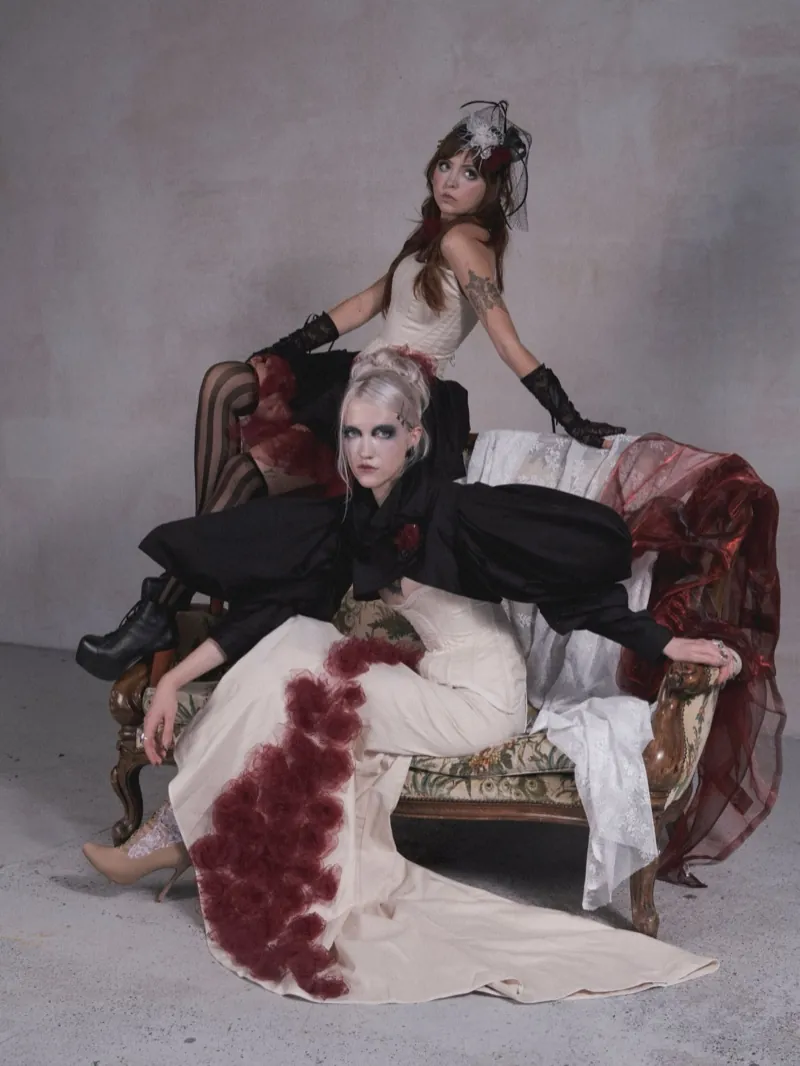 Two models on a sofa: a blonde in a black bolero sprawls in the foreground, behind her a brunette in black lace gloves turns her head